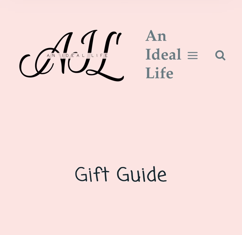 A soft pink webpage header features the logo “AIL” in a decorative script font, with the words “An Ideal Life” in small caps placed across the letters. To the right, the site title “An Ideal Life” is written in a clean, modern serif font, accompanied by a menu icon (three horizontal lines) and a magnifying glass symbol for search. Below, the title “Gift Guide” appears in a casual, handwritten-style font, indicating the section of the site focused on gift recommendations.