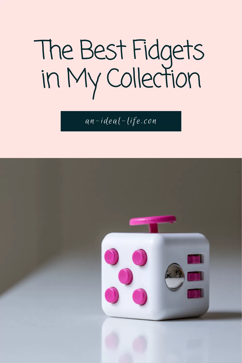 An image with text that reads, The Best Fidgets in My Collection” in a playful handwritten font, followed by the website an-ideal-life.com in a smaller, elegant script. The background is a soft pink color. Below the text is a photograph of a white fidget cube with pink buttons, a pink joystick, a silver rolling ball, and switches. The cube sits on a reflective white surface against a neutral background.