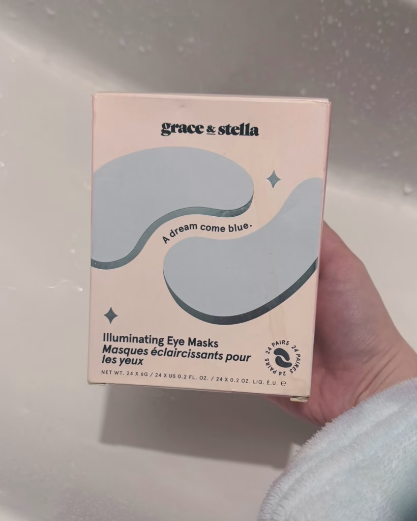 Hand holding a pastel pink box labeled “grace & stella” with blue under-eye patch graphics and text “A dream come blue. Illuminating Eye Masks.” 