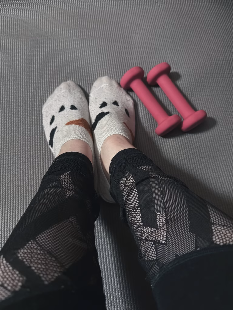A pair of feet wearing patterned socks, with black leggings, resting on a textured surface next to a pair of pink dumbbells.